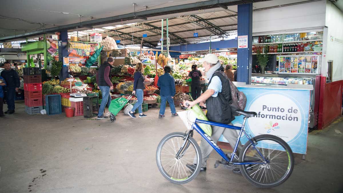 man walking his bike through a market with vegetable stands and people in the background