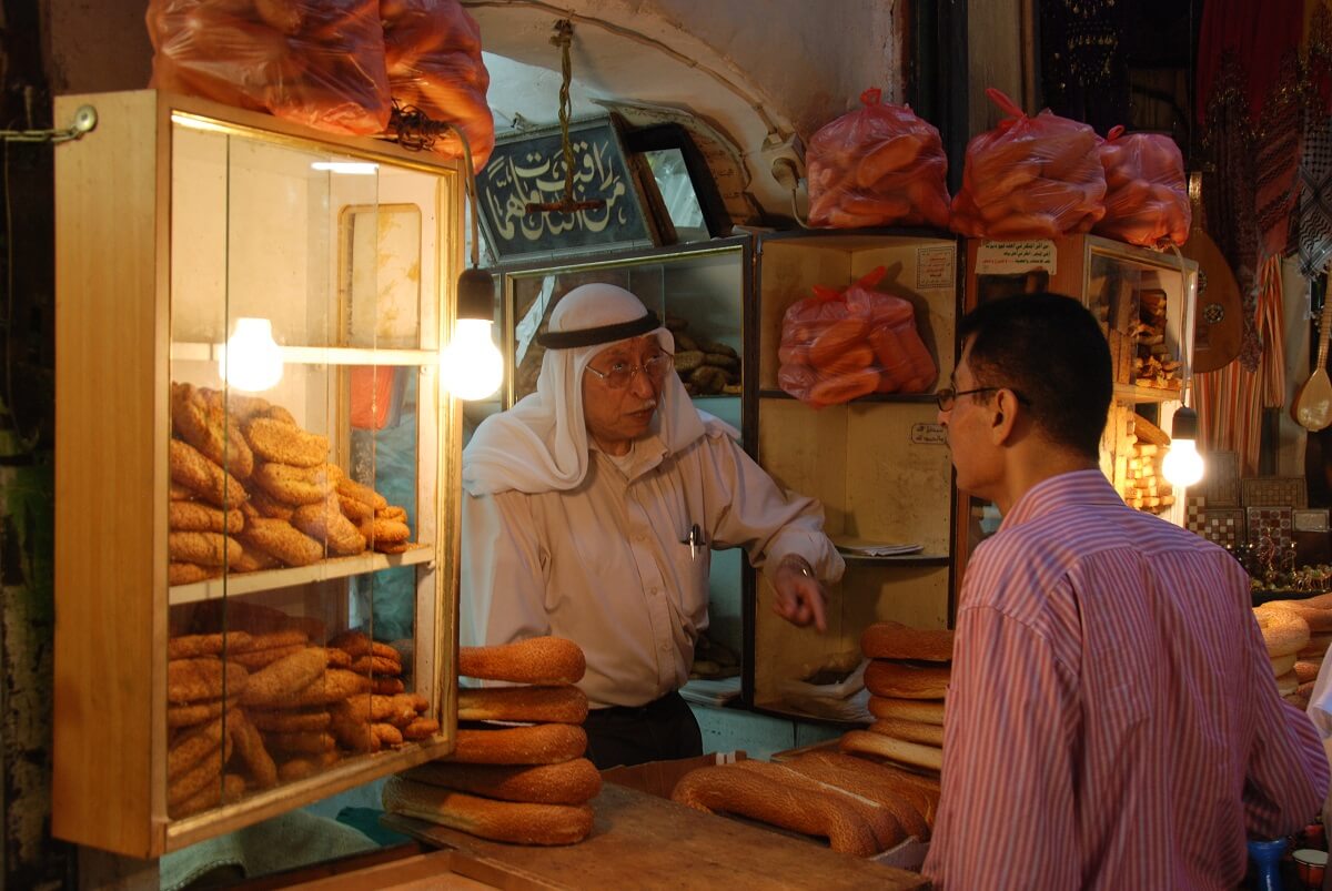 man purchasing bread from a market stall, speaking with another man.