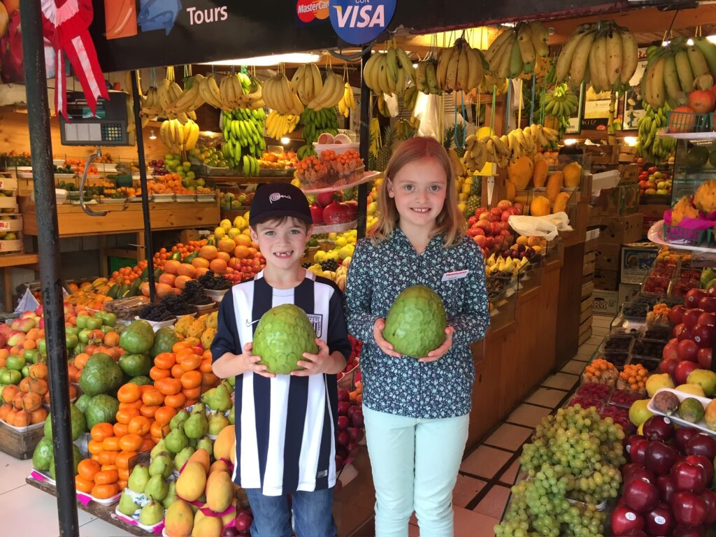 A boy and girl holding a melon in the aisle of an outdoor produce market stall.